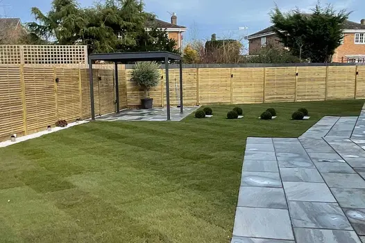 Proteus Grey aluminium pergola on paved area in far corner of lawn, bordered by fence and lawn. Built by Butt’s Landscapes.