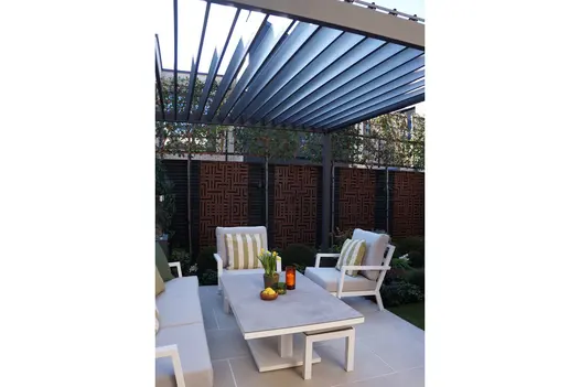 Proteus Grey metal pergola in urban back garden, louvred roof open, over outdoor furniture on patio with fence in background.