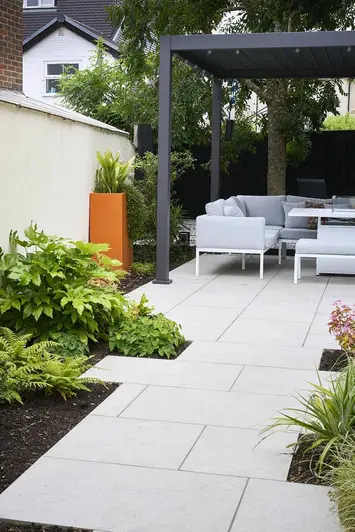 half-view of proteus grey aluminium pergola with furniture underneath, astor grey porcelain leads up with planting either side.