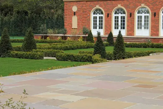 Angled shot of large brick house with a maze of hedges on a lawn, raj green indian sandstone paving shows off vast colours.