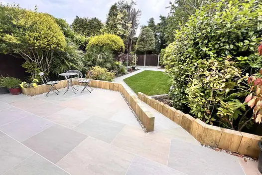 Slope of narrow path of raj green porcelain paving, from large patio, leads down to small lawn surrounded by large trees and bushes.