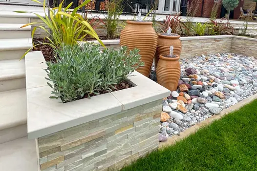 Water feature of 3 tall terracotta pots on pebbled area edged on 3 sides by stone-clad bed with Beige sawn sandstone coping.