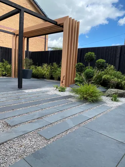 Brazilian Grey slate slabs and planks pave garden with pergola, dark fence and lushly planted narrow borders.