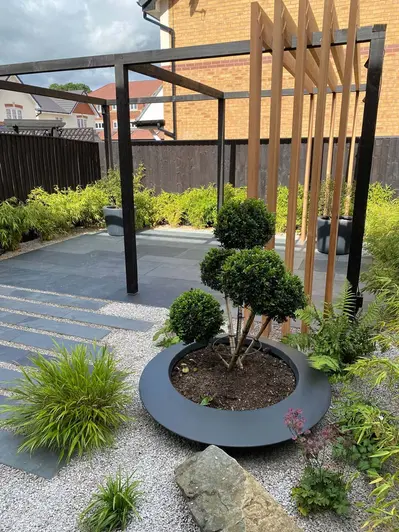 Fenced garden with pergola, paved with gravel and Brazilian Grey slate planks and slabs, bordered by lush planting.