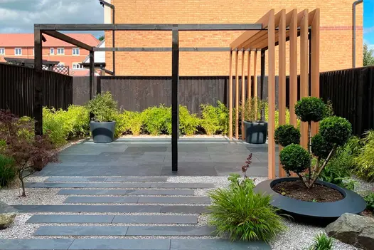 Dark-fenced garden paved with Brazilian Grey stone slabs  and planks set in gravel, with cloud-pruned shrub in shallow pot.