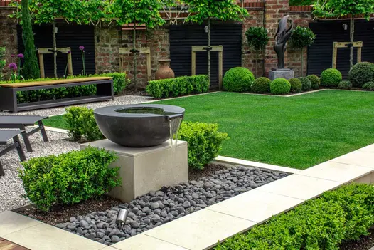 On plinth faced with Slab Khaki outdoor tiles, a round bowl water feature spills onto dark pebbles next to path of matching slabs.