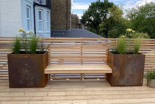 Wooden bench seat suspended between 2 corten steel tall trough planters on decked roof garden with slatted railings.