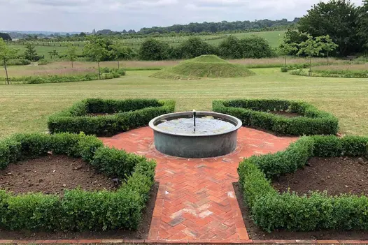 Paths of Romsey Antique clay pavers divide 4 shaped beds edged by low hedges that surround round water trough, with fields beyond.