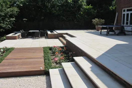 3 beige sawn sandstone bullnose steps lead down to pebbled area, away from beige patio, huge trees in the background.