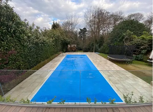 In mature garden, Frosty Grey Porcelain outdoor tiles, laid by S&D Paving, surround long, rectangular blue-lined swimming pool.
