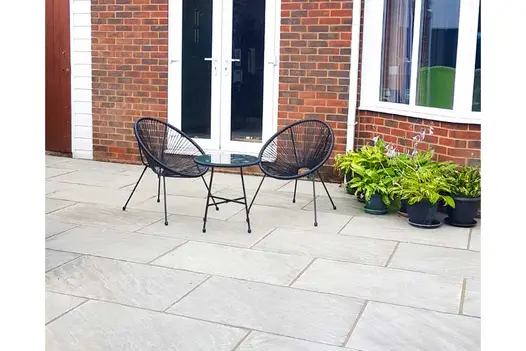2 wire bucket chairs and table sit on Kandla Grey sandstone paving outside house with french doors and potted ferns under window.