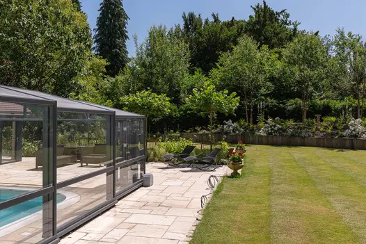 Tumbled Mint project pack sandstone paving patio with lawn on 2 sides, surrounds glass-covered swimming pool. Tall trees beyond.