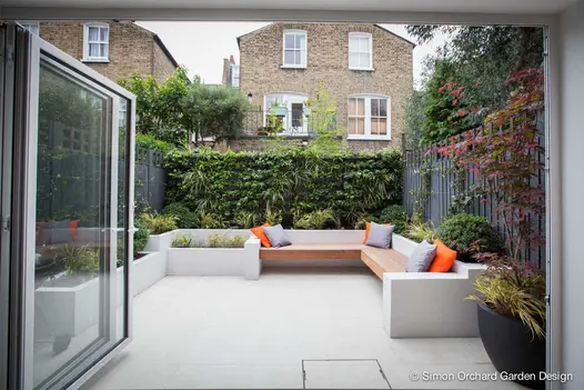 View through open bifold doors to small garden paved with Sandy White Porcelain. White raised beds. Wooden cantilevered bench.