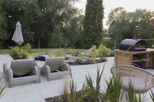 Sandy White Porcelain patio pierced with flowerbeds. Lawn and trees beyond. With outdoor kitchen and loungers. By Creative Roots.