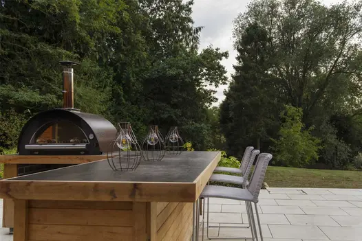 Wooden outdoor bar and table-top pizza oven with 3 stools, on Sandy White Vitrified Porcelain Paving, designed by Creative Roots.