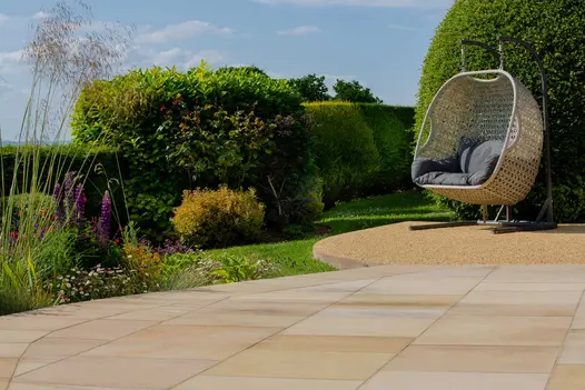 Rattan swing seat on gravel area next to large topiary and curved buff sawn paving slabs next to grassed and hedged border.