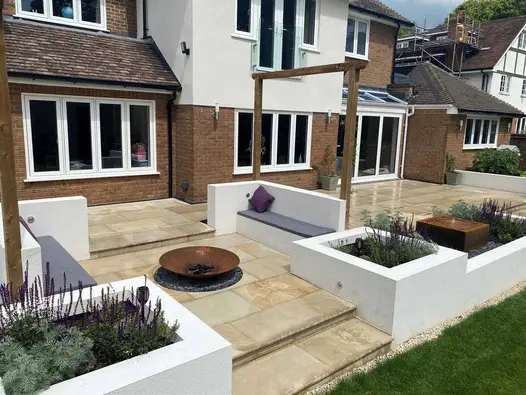 Light buff sawn sandstone patio across back of wide suburban house with Corten steel fire bowl between white inbuilt benches.