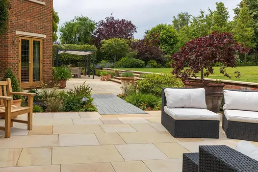Decking strip runs between 2 sections of Buff Smooth sandstone patio separated by planted beds and edged by lawn’s retaining wall.