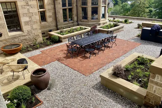 Gravel-paved courtyard of stone house, with rectangle of Romsey Antique Belgian bricks under large metal dining set.