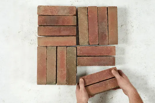 2 hands complete a square by adding 2 Seville clay pavers to 14 others laid in 4 blocks at right angles to each other. 