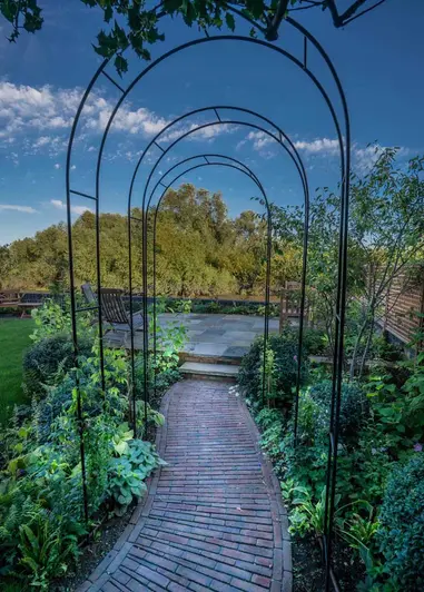Path of Abbey Dark Multi clay paving curves slightly under 3 metal rose arches, leading to 2 steps up to patio with wooden seats.