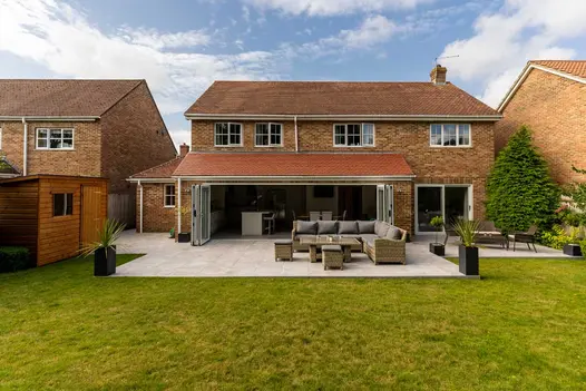Rear of suburban house with wide bifold doors open onto Silver Contro Porcelain patio with black planters and rattan furniture.
