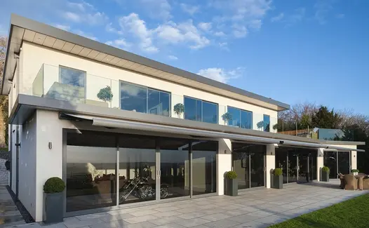 Plain patio of Silver Grey granite paving runs along back of modern house with large windows and  house-width awning.