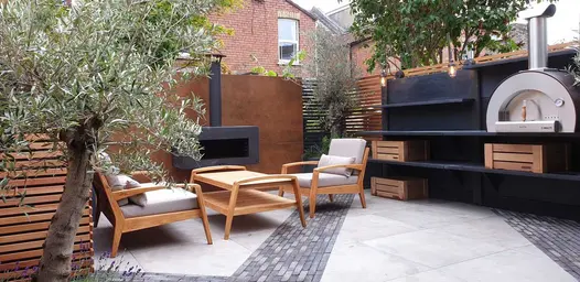 Corner of urban garden paved with porcelain and wide stripes of Silver Grey Multi clay pavers. Lounge set and pizza oven on shelves.