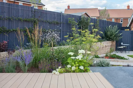Small deck of mocha designboard runs along raised and ground level flowerbeds leading down to pebble and lawn area.