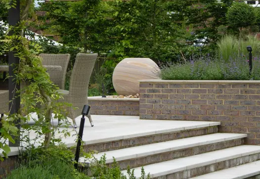 4 Jura Beige sawn limestone steps with brick risers rise to patio with outdoor furniture and spherical water feature on pebbles.