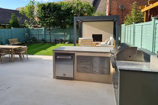 Outdoor kitchen in green-fenced garden, with pergola and wooden dining set on Slab Khaki porcelain outdoor tiles, with lawn beyond.