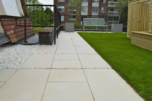 Path of Slab Khaki porcelain outdoor tiles leads to wooden bench and planters, between black railings and grassy border. 