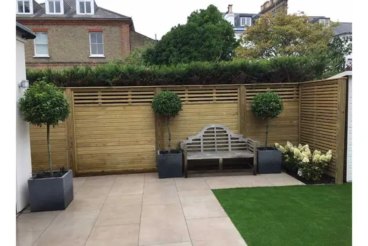 Low maintenance rear garden paved in Slab Khaki porcelain paving with a small area of artificial grass. Decorated with pots and a bench.