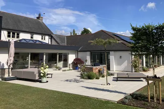 Square patio of slab khaki porcelain outdoor tiles UK laid in angle between house and extension. Glass doors open onto it on 2 sides.