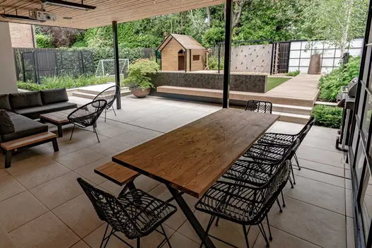 View across garden from patio paved with Slab Khaki porcelain slabs under pergola. Decking path leads to children’s play area.