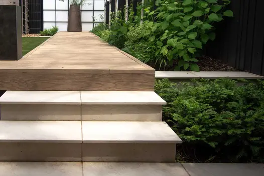 2 steps rise to decking walkway along side of planted bed. Slab khaki porcelain coping, with 40mm downstand, tops wall to right.