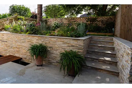 Silver grey porcelain slabs for steps rise between stone-clad retaining walls from terrace to decked area under mature trees.