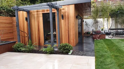 Cedar clad garden office with dark composite decking surround, natural lawn and a porcelain patio.