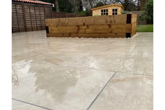 Large Ash Beige porcelain patio with light grey grouting lines wrapped around a large raised flower bed constructed from new wooden sleepers.