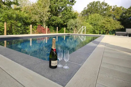 Champagne bottle and 2 glasses sit on Urban Grey porcelain swimming pool coping in midst of Smoked Oak Millboard decking.