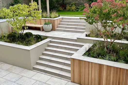 Steps flanked by trees in beds rise in 2 consecutive flights from paving to upper garden level. Jura Grey smooth coping caps walls.