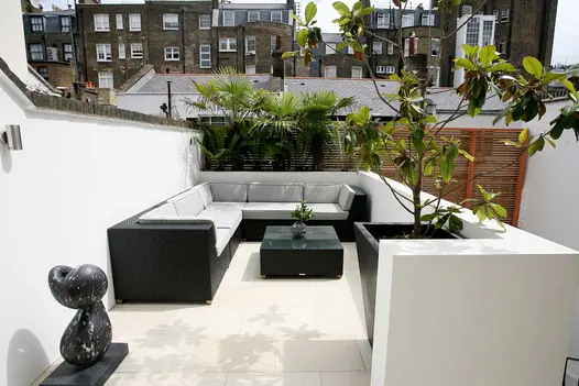 Black metal sculpture sits at entrance to seating area of Beige smooth sandstone paving bounded by low white wall on 2 sides.
