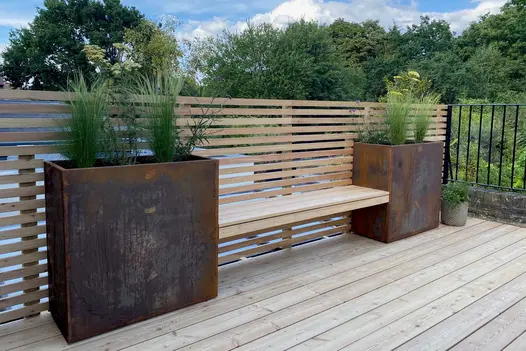 Treetops behind slatted fence which backs wooden bench seat supported on either side by Corten steel tall trough planters.