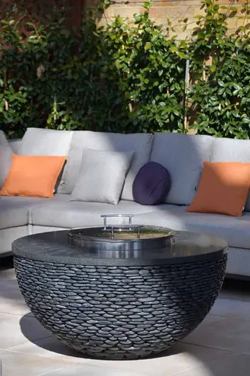 Bowl-shaped dark-coloured fire pit with stainless steel cover sits on Jura Beige contemporary limestone paving next to sofa.
