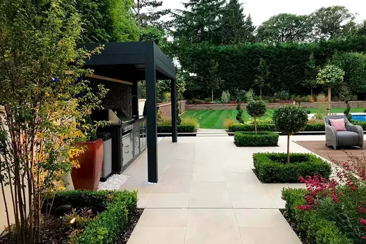 Golden Stone Porcelain patio with outdoor kitchen, metal pergola and decking set flush to paving, by The Landscape Design Studio.