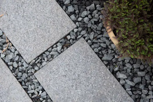 Close-up of Dark Grey granite plank paving, laid with wide joints of matching gravel, next to evergreen in terracotta pot.