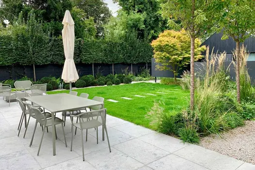 Large Jura Grey sawn limestone patio next to lawn with plank stepping stones, topiary balls, and grasses planted between trees.
