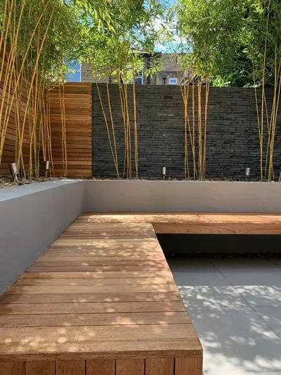 L-shaped wooden bench floats around white rendered walls with built in flowerbeds, black slate cladding with trees in the background.