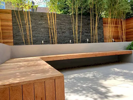 Wooden bench on light patio lines raised flower bed with black slate clad wall and new trees behind.