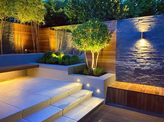 View at night see steps lead up to patio area from decking, black slate cladding used on wall with wooden clad fencing and a mirror.